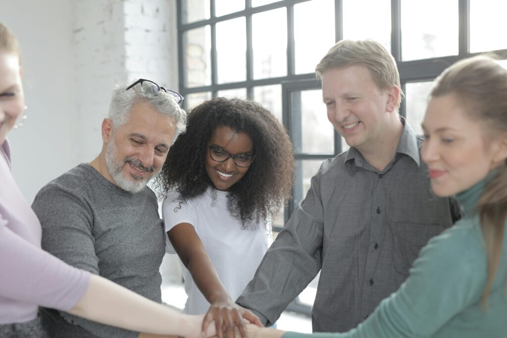 Cheerful multiracial coworkers in casual wear uniting hands while standing behind brick wall near big fenced window and looking down in sunlight