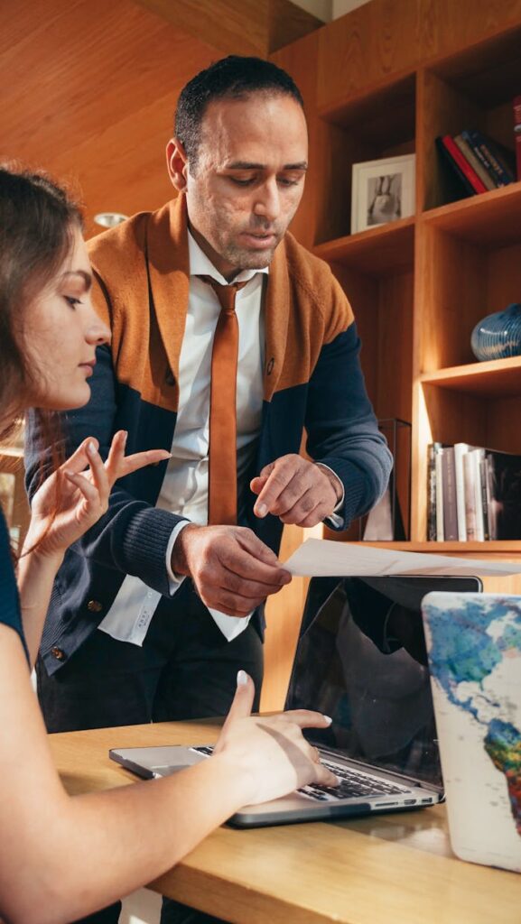 Two colleagues discussing work over a laptop in a modern office space with bookshelves in the background.