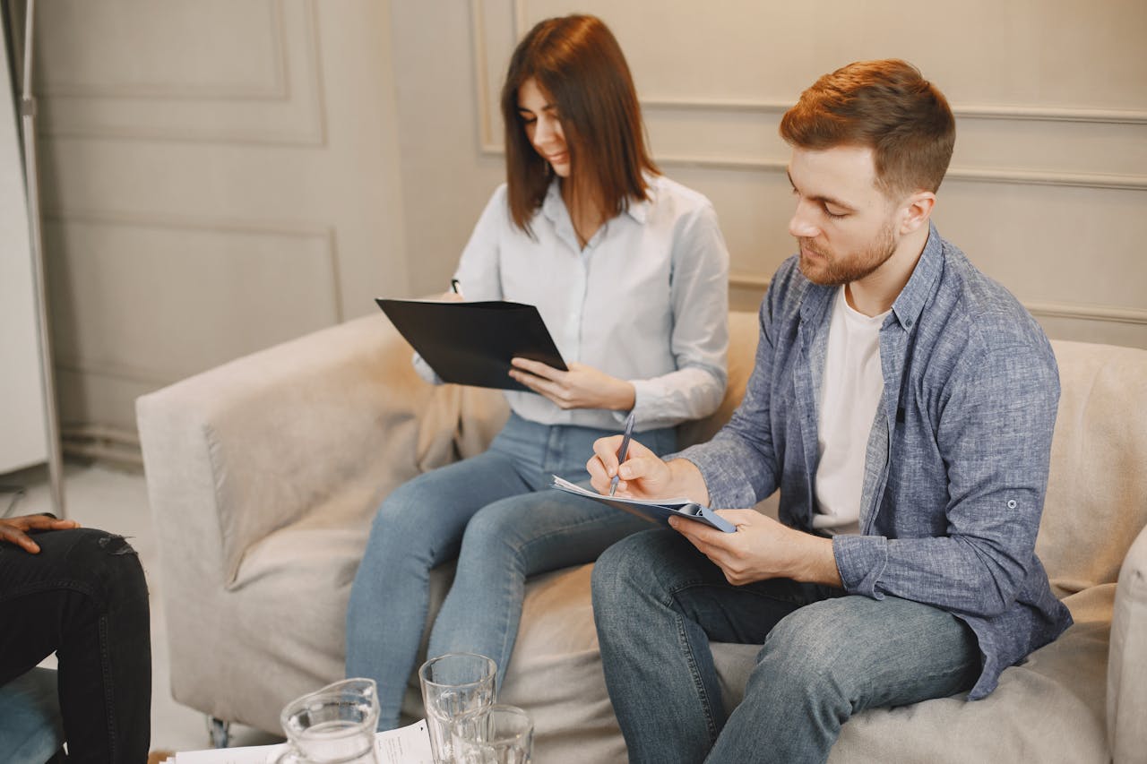 A young couple sitting on a sofa, writing and examining documents in a cozy indoor setting.